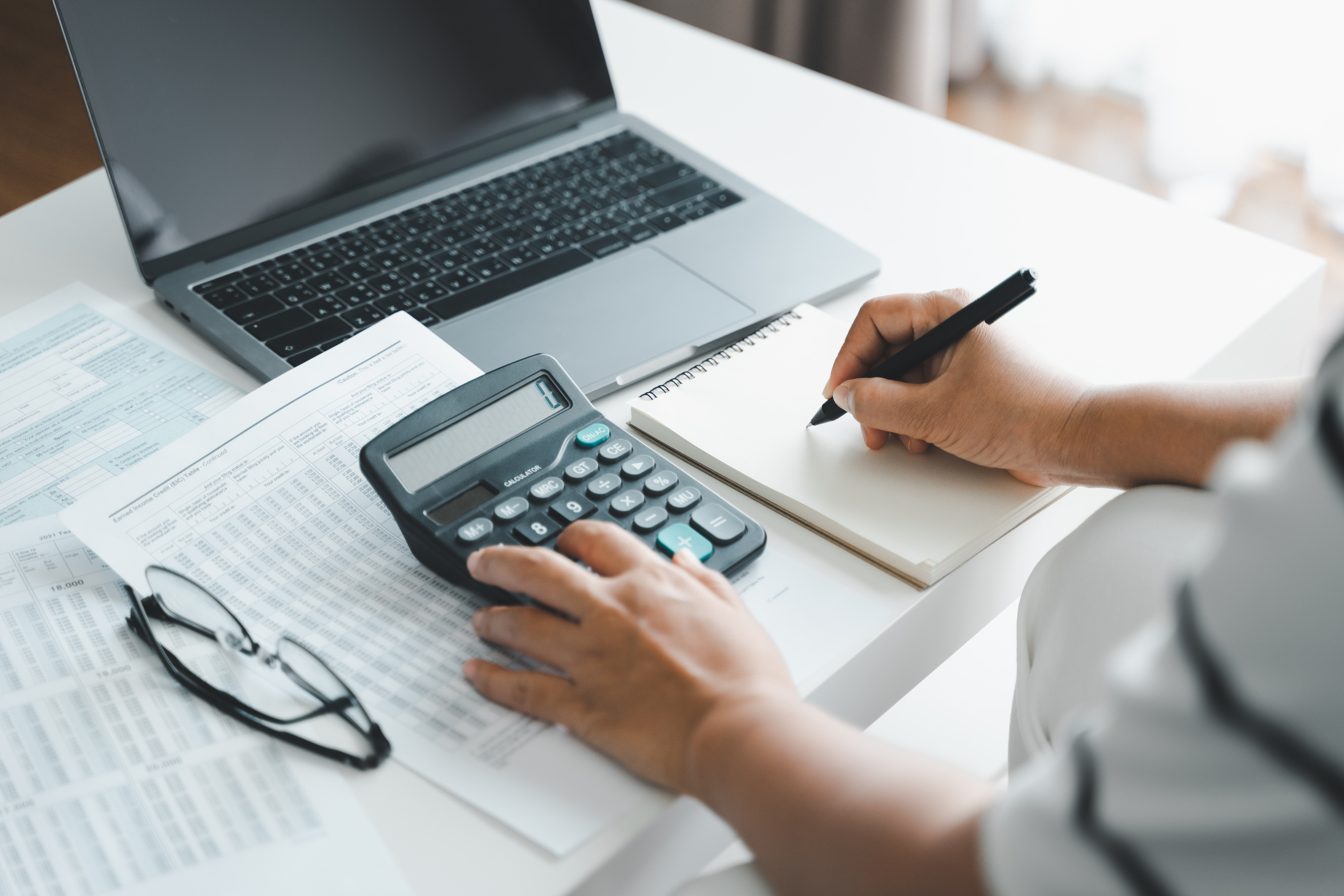 Person using a calculator and taking notes beside tax forms and a laptop